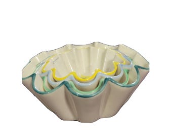May include: A set of four nested ceramic bowls. The bowls are off-white with scalloped edges and wavy, colorful trim in yellow, blue, and green. The bowls are designed to nest inside each other for storage.