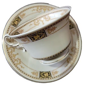 May include: A white teacup and saucer set with gold and black decorative trim. The teacup has a curved handle and sits on a matching saucer. The set features a detailed floral and geometric pattern.