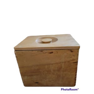 May include: A square wooden box with a light tan finish and visible natural wood grain patterns. The box features a flat, removable lid with a centered circular knob handle. The sides of the box are joined with visible stitching or lacing.