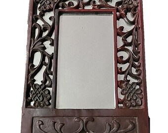 May include: A rectangular, dark brown wooden mirror with intricate floral and scroll carvings. The frame features a central mirror surrounded by detailed openwork designs and decorative elements. The mirror is supported by a base with curved accents.