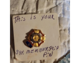 Könnte beinhalten: Eine goldfarbene Mitgliedsnadel mit roten, weißen und blauen Akzenten. Die Nadel befindet sich auf einer weißen Karte mit handschriftlichem Text: "This is your SYR MEMBERSHIP PIN."