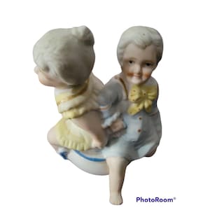 May include: A porcelain figurine of two children, a girl in a yellow dress and a boy in blue overalls, riding a white and blue porcelain ball.