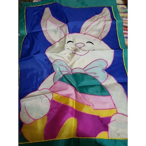 May include: Easter-themed flag featuring a smiling white bunny holding a colorful egg. The bunny has pink accents on its ears and a teal bow. The background is blue with green borders, creating a festive and cheerful design.