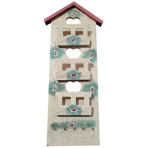 May include: A decorative wooden wall organizer shaped like a house. It features a red roof, heart-shaped cutouts, and rectangular openings. The off-white surface is adorned with green and pink floral accents, and has a distressed, crackled finish.