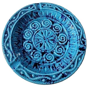 May include: A blue ceramic ashtray with a decorative design of swirls and a splatter pattern. The ashtray has a raised rim and a center depression.