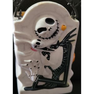 May include: A white Halloween-themed container featuring Jack Skellington from The Nightmare Before Christmas. The container has a tombstone shape with a black handle and a black skeleton figure. A small orange pumpkin is also visible.