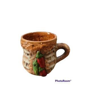 May include: A brown ceramic mug with a textured surface and a handle. The mug has a green and red chili pepper design.