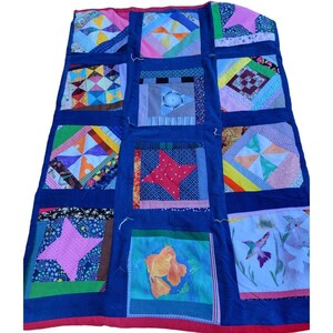 May include: A handmade quilt with a patchwork design. The quilt features a dark blue background with various colorful squares, each showcasing unique patterns and images, including stars, geometric shapes, and a goldfish. The quilt is trimmed with a red border.