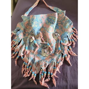 May include: A handmade, bohemian-style bag with a paisley pattern in shades of pink, blue, and yellow. The bag features beaded fringe in pink, blue, and yellow, and a strap for carrying. The bag has a unique, artistic design.
