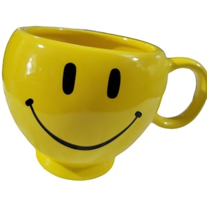 May include: Yellow ceramic mug shaped like a smiley face with black eyes and a black smile.