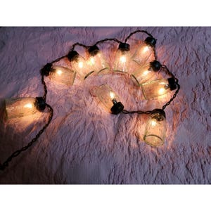May include: A string of decorative lights featuring small, clear glass jar-shaped bulbs with black caps. The lights are illuminated, casting a warm, yellow glow. The string is arranged on a textured, pink surface.