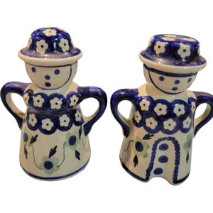 May include: A pair of ceramic salt and pepper shakers shaped like women wearing blue and white dresses with floral patterns. The shakers have handles and are decorated with blue and white flowers.