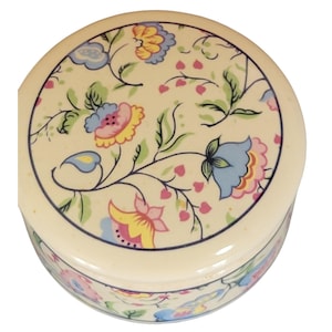May include: A round, cream-colored ceramic trinket box with a floral design. The lid and sides feature a pattern of blue, pink, yellow, and green flowers and leaves. A dark blue line borders the lid.