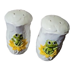 May include: A set of two white ceramic salt and pepper shakers with green frog and yellow flower designs.