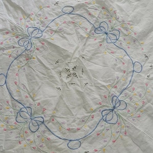 K&ouml;nnte beinhalten: Wei&szlig;e Tischdecke mit einem zarten floralen Stickmuster. Die Stickerei zeigt blaue Schleifen, gr&uuml;ne Stiele und rosa, gelbe und lila Bl&uuml;ten.