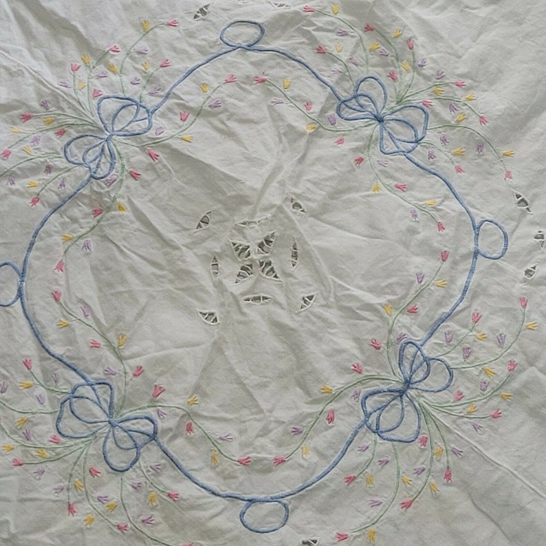 K&ouml;nnte beinhalten: Wei&szlig;e Tischdecke mit einem zarten floralen Stickmuster. Die Stickerei zeigt blaue Schleifen, gr&uuml;ne Stiele und rosa, gelbe und lila Bl&uuml;ten.