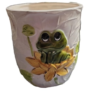 May include: A white ceramic mug featuring a raised, three-dimensional design of a green frog sitting inside a yellow lotus flower. The mug surface has a textured leaf pattern, and the frog has large, dark eyes looking upward with a subtle smile.