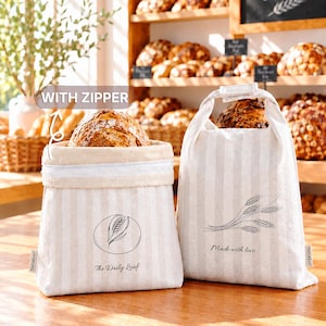 May include: Two beige and white striped fabric bread bags with handles. The left bag features a zipper, a wheat stalk graphic, and the text "The Daily Loaf." The right bag displays a wheat stalk illustration and the text "Made with love."