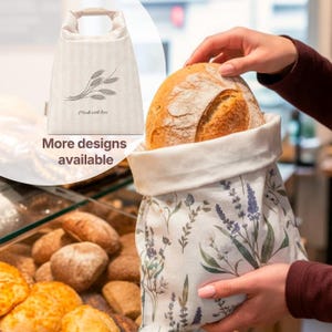 May include: A fabric bread bag featuring a botanical print of lavender and wildflowers, holding a round loaf of crusty bread. A smaller inset shows a beige bag with a wheat stalk graphic and the text "Made with love" printed on it.