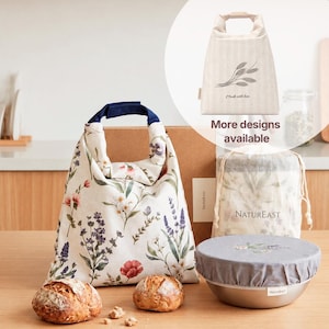 May include: A cream-colored fabric lunch bag with a floral pattern featuring lavender, red, and white flowers, paired with a dark blue handle. Beside it sits a metal bowl covered with a gray fabric lid and a drawstring bag labeled NatureEast.
