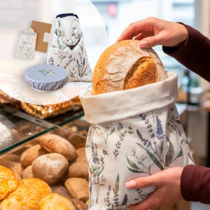 May include: A reusable fabric bread bag featuring a botanical print of lavender and wildflowers, containing a round loaf of crusty bread. The set includes a matching fabric bowl cover and a small rectangular sachet with printed text.