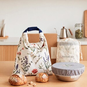 May include: A cream-colored fabric lunch bag with a floral print and dark blue handle, a matching drawstring pouch labeled NATUREAST, and a bowl covered with a gray fabric lid featuring a small floral design, alongside two round crusty bread loaves.