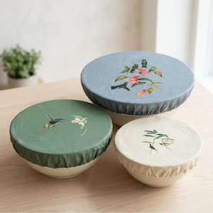 May include: Three ceramic bowls with fabric covers. The covers are in shades of green, blue, and cream, each adorned with embroidered floral and hummingbird designs. The bowls are on a light wood surface.