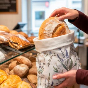 May include: A white fabric bread bag featuring a botanical print of purple lavender and green foliage, containing a round, golden-brown crusty loaf of bread. The bag has a folded top edge and is held open to reveal the baked bread inside.