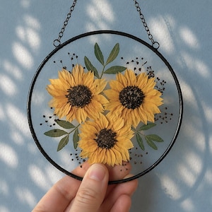 Op de afbeelding: Ronde wanddecoratie met een zwart frame, met drie gedroogde zonnebloemen met gele bloemblaadjes en donkerbruine centra. Groene bladeren en kleine takjes omringen de bloemen. Opgehangen aan een fijne ketting.