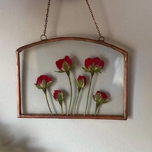 Könnte beinhalten: Ein rechteckiger Wandbehang aus Glas mit einem Kupferrahmen und einer dünnen Kupferkette zum Aufhängen. Zwischen den beiden klaren Glasscheiben befinden sich fünf gepresste rote Rosenknospen mit grünen Stielen und Blättern.
