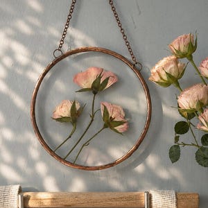 May include: A circular copper-toned metal frame hangs by a chain, enclosing three pressed pink roses between two glass panes. To the right, a ceramic vase holds a bouquet of fresh pink roses. A textured beige fabric hangs from a wooden rod below.