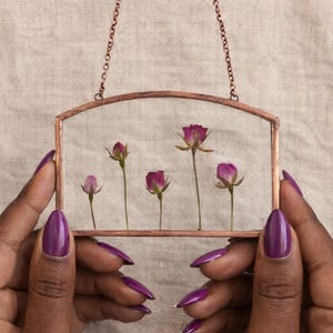 May include: A rectangular glass frame with a copper metal border and a curved top edge, suspended by a thin copper chain. Inside the clear glass are five pressed pink rosebuds with green stems, arranged in a row of varying heights.