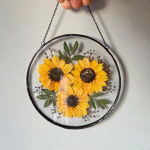 Op de afbeelding: Ronde glazen wanddecoratie met drie geperste gele zonnebloemen met donkerbruine centra. De bloemen zijn omgeven door groene bladeren en kleine bruine knoppen, ingekapseld in een zwart metalen frame en opgehangen aan een ketting.