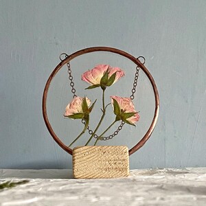 May include: A circular copper frame holds three pressed pink roses suspended by thin metal chains. The frame is mounted on a rectangular, light-colored wooden base with a visible grain texture. The flowers feature soft pink petals.