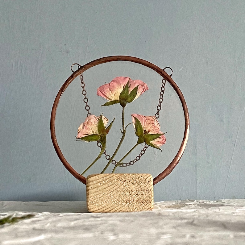 May include: A circular copper frame holds three pressed pink roses suspended by thin metal chains. The frame is mounted on a rectangular, light-colored wooden base with a visible grain texture. The flowers feature soft pink petals.