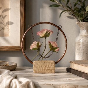 May include: A circular copper-toned metal frame mounted on a rectangular textured stone base. Three dried pink roses are suspended inside the ring by thin metal chains. The frame features a metallic finish and a simple, clean structural design.
