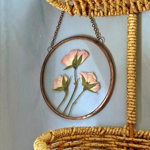May include: A circular wall hanging featuring three pressed light pink rosebuds encased between two clear glass panes within a thin copper frame. The piece is suspended by a delicate copper-toned metal chain attached to the top of the frame.