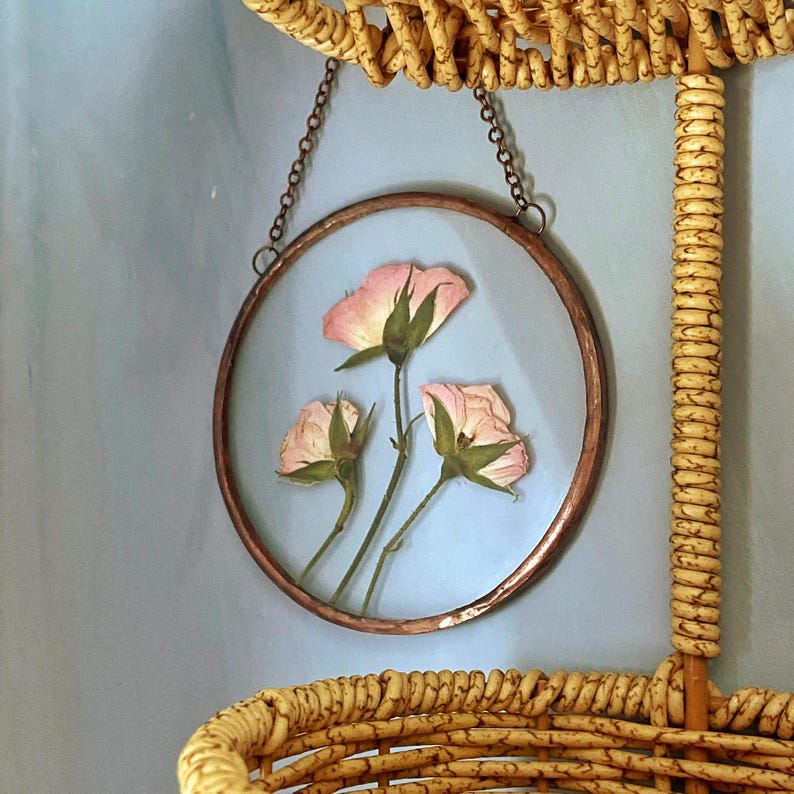 May include: A circular wall hanging featuring three pressed light pink rosebuds encased between two clear glass panes within a thin copper frame. The piece is suspended by a delicate copper-toned metal chain attached to the top of the frame.