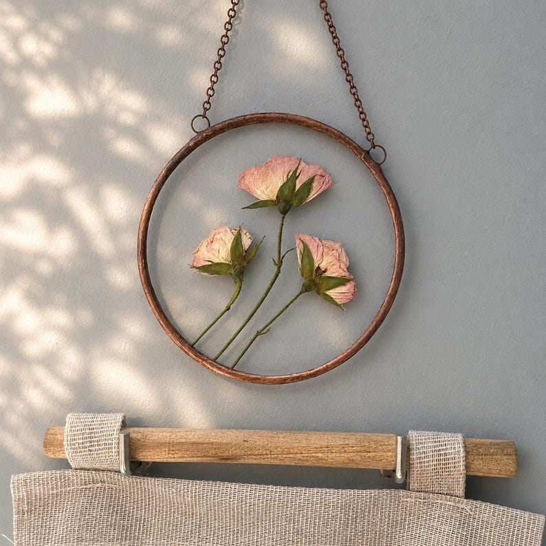 May include: A circular copper-toned metal frame containing three pressed pink rosebuds with green stems, suspended by a matching copper-colored chain. The frame is positioned above a light beige textured fabric hanging from a wooden rod.