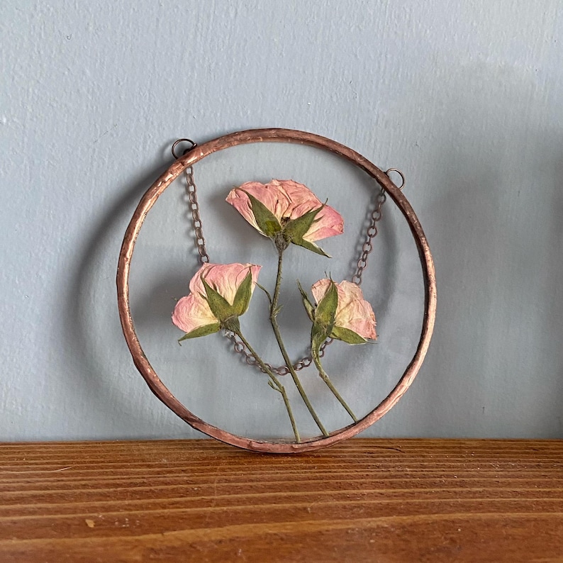 May include: A circular wall hanging featuring three pressed pink roses encased between two clear glass panes. The glass is framed by a thin copper border with a metallic finish. A delicate metal chain is attached to the top for hanging purposes.