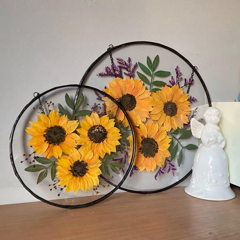 Op de afbeelding: Twee ronde wanddecoraties van helder glas met geperste zonnebloemen, groene bladeren en paarse accenten. De zonnebloemen zijn heldergeel met donkerbruine centra. De frames zijn van zwart metaal met ophangkettingen. Rechts staat een wit keramieken engelenbeeldje.