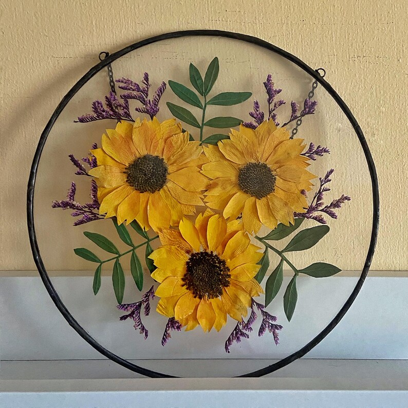 May include: A round, clear glass piece with a black metal frame, showcasing a floral arrangement. The arrangement features three bright yellow sunflowers with dark brown centers, surrounded by green leaves and purple sprigs. A chain is attached for hanging.