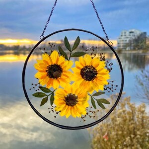 Op de afbeelding: Ronde, heldere glazen hangdecoratie met drie felgele zonnebloemen en groene bladeren. De bloemen hebben donkerbruine centra. De decoratie hangt aan een zwarte metalen ketting. De achtergrond toont een meer en een bewolkte lucht.
