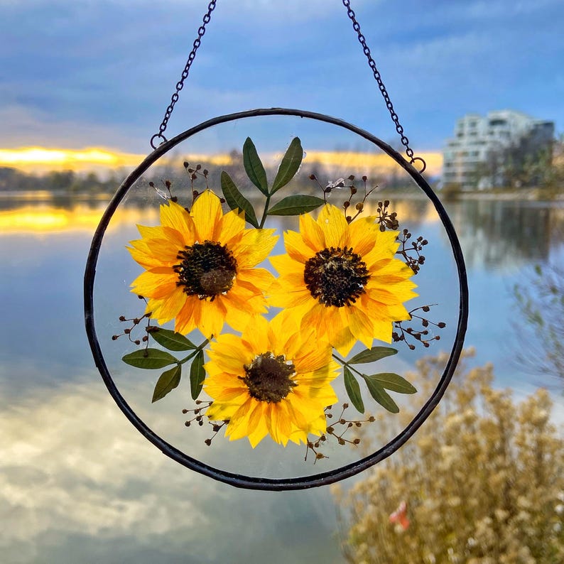 Op de afbeelding: Ronde, heldere glazen hangdecoratie met drie felgele zonnebloemen en groene bladeren. De bloemen hebben donkerbruine centra. De decoratie hangt aan een zwarte metalen ketting. De achtergrond toont een meer en een bewolkte lucht.