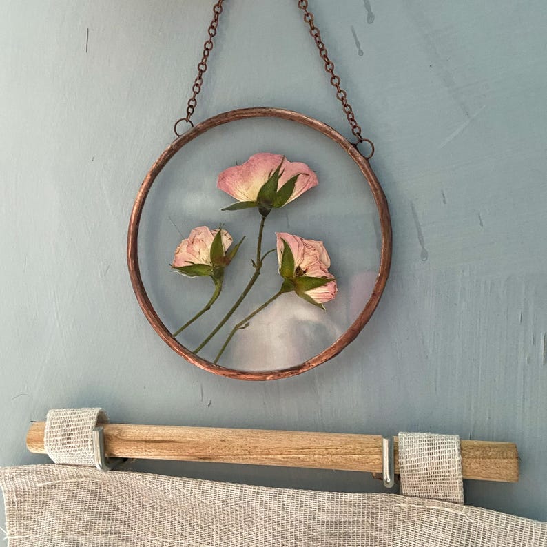 May include: A circular wall hanging featuring three pressed pink roses encased between two clear glass panes within a copper-toned metal frame. The piece is suspended by a matching copper-toned metal chain attached to the top of the circular frame.