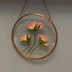 May include: A circular wall hanging featuring three pressed pink rosebuds encased between two clear glass panes. The glass is framed in a thin copper metal border and suspended by a matching copper-colored metal chain for display purposes.