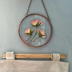 May include: A circular wall hanging featuring three pressed pink rose blossoms encased between two clear glass panes within a copper-toned metal frame, suspended by a matching copper-toned metal chain for display on a vertical surface.