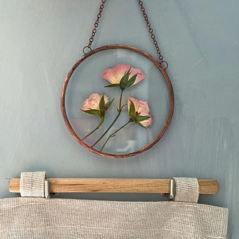 May include: A circular wall hanging featuring three pressed pink rose blossoms encased between two clear glass panes within a copper-toned metal frame, suspended by a matching copper-toned metal chain for display on a vertical surface.