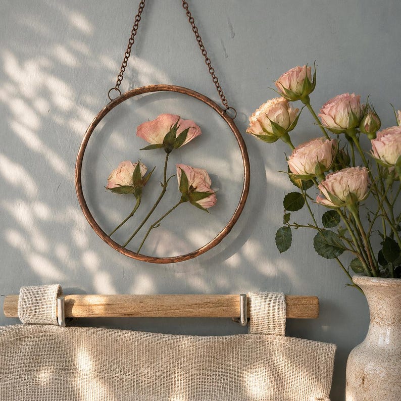 May include: A circular copper-toned metal frame hangs by a chain, enclosing three pressed pink roses between two glass panes. To the right, a ceramic vase holds a bouquet of fresh pink roses. A textured beige fabric hangs from a wooden rod below.