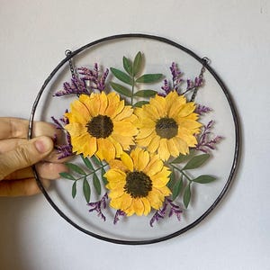 Op de afbeelding: Ronde, heldere glazen wanddecoratie met drie gele zonnebloemen met donkerbruine centra. Groene bladeren en paarse takjes omringen de bloemen, alles in een zwart metalen frame. Een ketting is bevestigd om op te hangen.